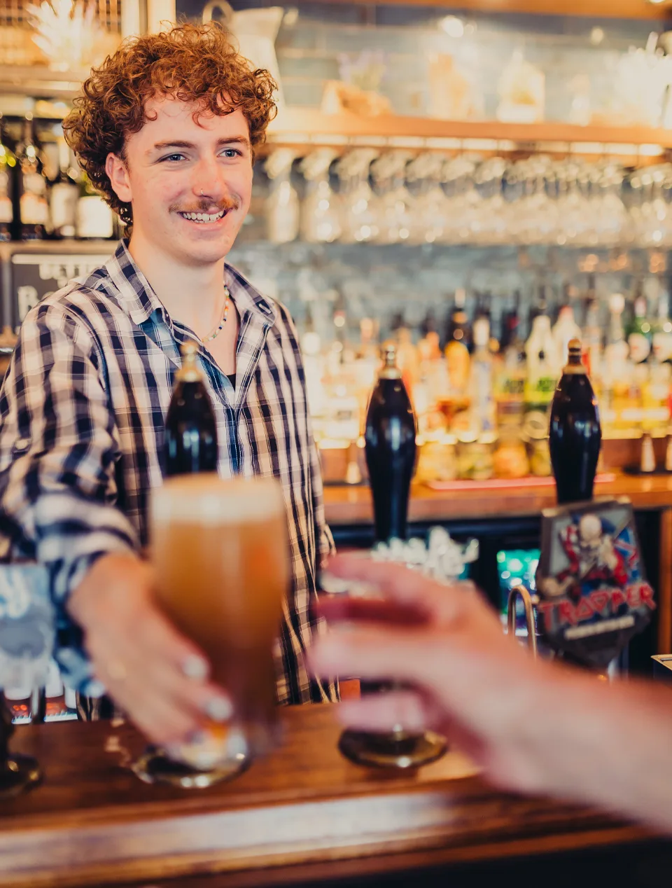 A bartender behind a pub bar hands a pint of beer to a customer. Taps, bottles and rows of hanging glasses are visible in the background under warm lighting.